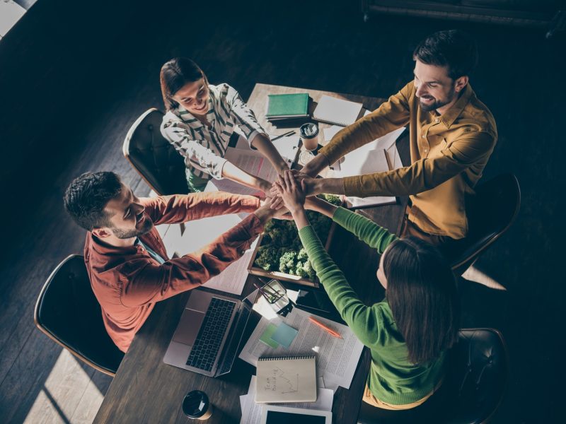 Top above high angle view of four nice cheerful professional businesspeople wearing casual formalwear putting palms together over desktop at modern industrial loft style interior work place station