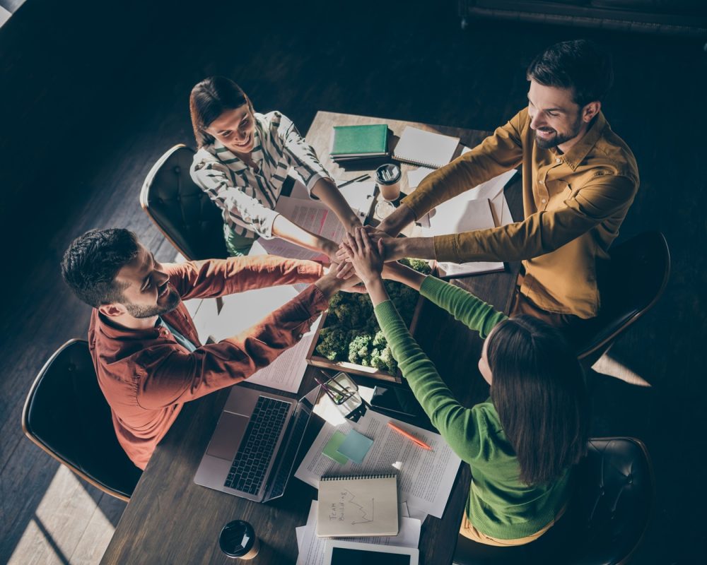 Top above high angle view of four nice cheerful professional businesspeople wearing casual formalwear putting palms together over desktop at modern industrial loft style interior work place station